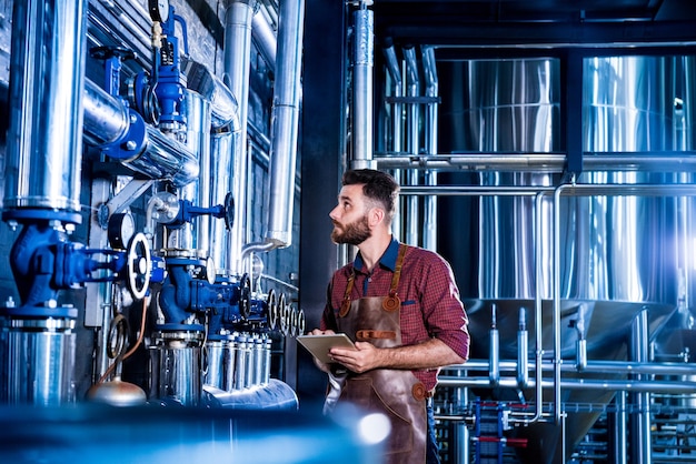 Junger männlicher Brauer in Lederschürze, der den Prozess der Biergärung in einem modernen Brauereibetrieb überwacht.