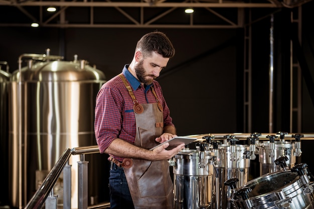 Junger männlicher Brauer in Lederschürze, der den Prozess der Biergärung in einem modernen Brauereibetrieb überwacht.