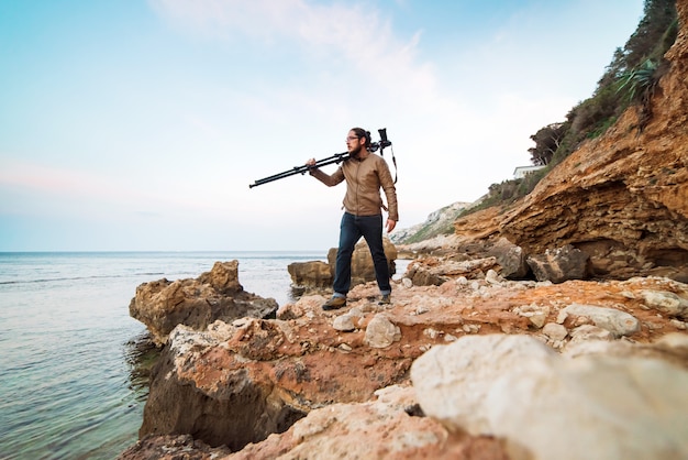 Junger männlicher Fotograf, der auf Felsenstrand mit Kamera auf Stativ geht