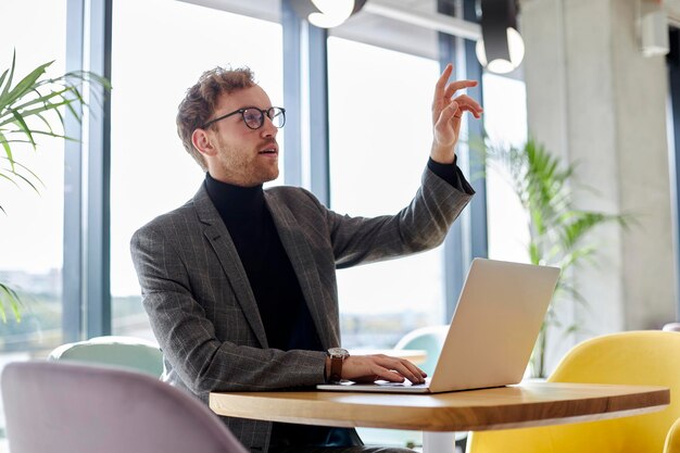 Junger männlicher Freiberufler, der jemanden begrüßt, der am Schreibtisch sitzt und Online-Arbeit auf dem Laptop sicherstellt