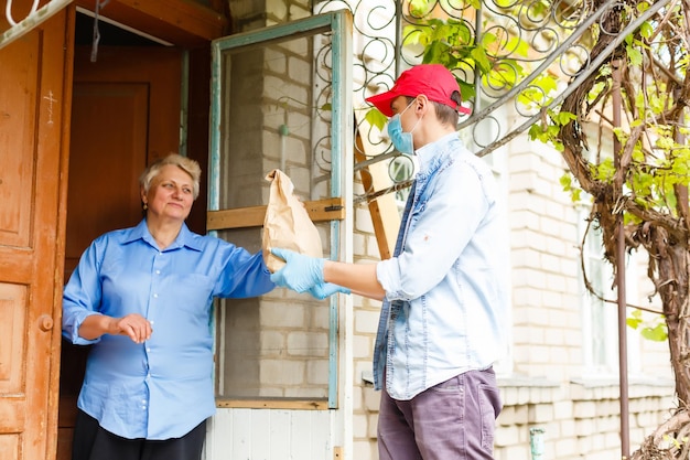 Junger männlicher Freiwilliger in Maske gibt einer älteren Frau Kisten mit Essen in der Nähe ihres Hauses. Sohnmann hilft einer alleinerziehenden älteren Mutter. Familienunterstützung, Fürsorge. Quarantäne, isoliert. Coronavirus (COVID-19. Spende