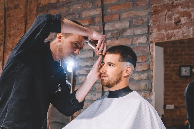 Junger männlicher Friseur rasiert den Kopf des Kunden mit einem elektrischen Trimmer. Junger bärtiger Mann, der sich vom Friseur im Friseursalon die Haare schneiden lässt. Werbe- und Friseurkonzept