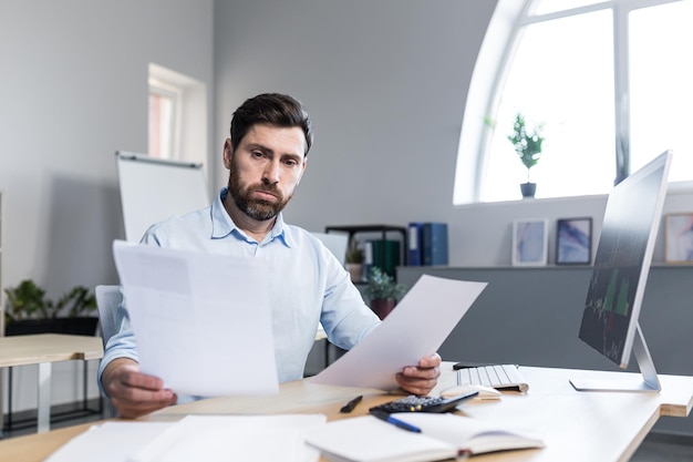 Junger männlicher Geschäftsmann Buchhalter, Ökonom, Finanzier, arbeitet mit Dokumenten, Papieren, Berichten, Zeitplänen, die an seinem Schreibtisch am Computer sitzen