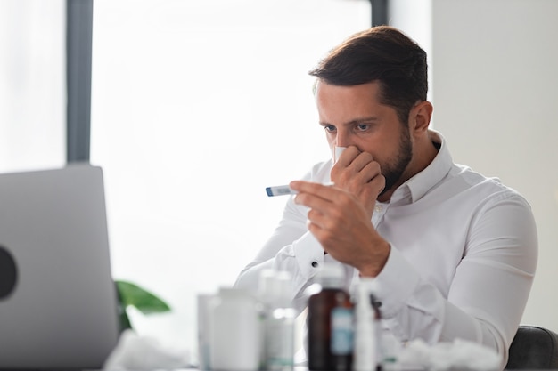 Junger männlicher geschäftsmann putzt sich die nase und misst die temperatur mit einem thermometer, während er im büro sitzt, grippevirus, erkältung, unwohlsein konzept