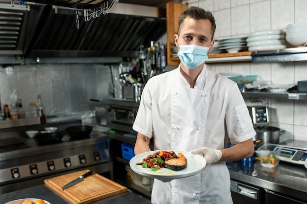 Junger männlicher Koch in weißer Uniform und Schutzmaske, der Teller mit gebratenem Lachs und Gemüse hält, während er vor der Kamera steht