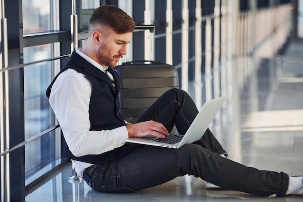 Junger männlicher Passagier in eleganter formeller Kleidung sitzt mit Laptop in der Flughafenhalle.