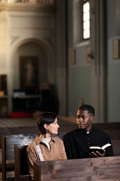 Junger männlicher Priester und Frau unterhalten sich in der Kirche, während sie eine Bibel halten