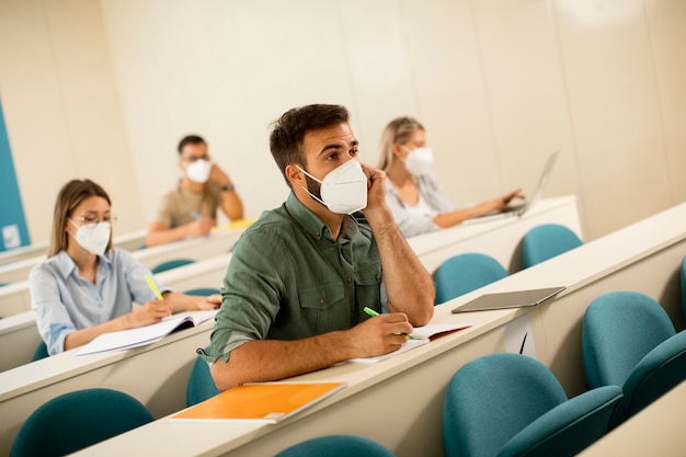 Junger männlicher Student, der medizinische Gesichtsschutzmaske für Virenschutz am Hörsaal trägt