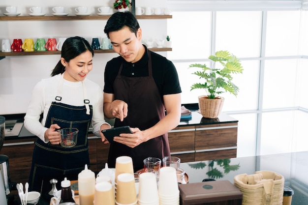 Junger männlicher und weiblicher Barista-Cafébesitzer, der innerhalb der Kaffeetheke steht und über Kundenbestellung von einer Tablette mit Lächeln spricht.