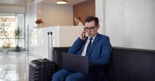 Junger männlicher Unternehmer sitzt mit Gepäck in der Hotellobby und telefoniert