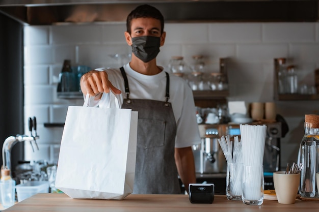 Junger Mann Barista mit Gesichtsmaske und Schürze, der dem Kunden im Café ein POS-Terminal gibt