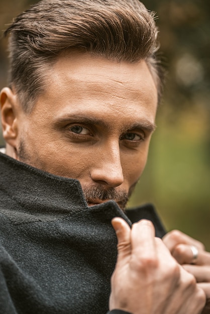 Junger Mann bedeckt sein Gesicht mit einer Ecke seines Mantels, der auf der Straße in einem herbstlichen Herbstmantel steht