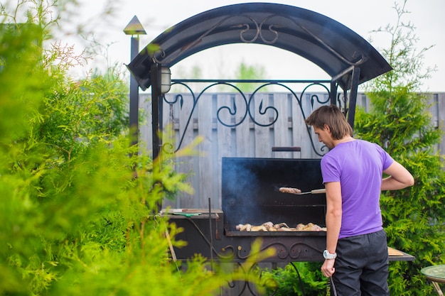 Junger Mann brät Steaks auf dem Grill, der in seinem Yard im Freien ist