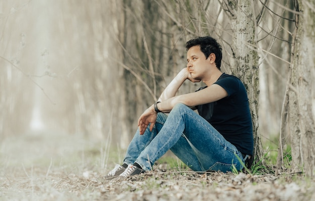 junger Mann, der an einem Baum im Wald sitzt und über das Leben nachdenkt und sich ausruht.