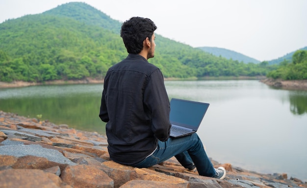 Junger Mann, der an einem Laptop arbeitet und in der Natur sitzt Workation und Digital Nomad Konzept
