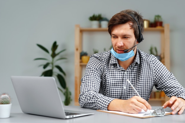 Junger Mann, der an Laptop mit Headset und Gesichtsmaske zu Hause arbeitet