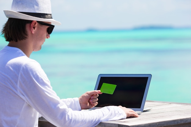 Junger Mann, der an Laptop mit Kreditkarte am tropischen Strand arbeitet