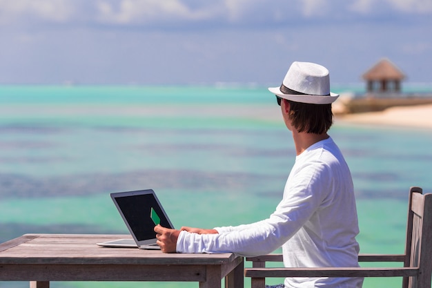 Junger Mann, der an Laptop mit Kreditkarte am tropischen Strand arbeitet