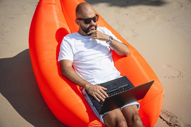 Junger Mann, der an seinem Laptop am Strand arbeitet, während er auf einem aufblasbaren Sofa sitzt