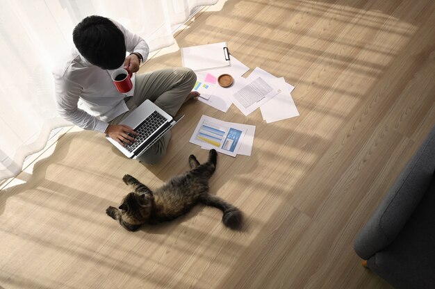 Junger Mann, der an seinem Laptop mit seiner Katze in bequemem Zuhause arbeitet.