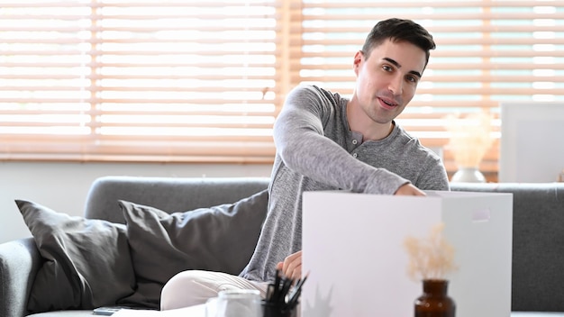 Junger Mann, der auf der Couch sitzt und Habseligkeiten in Kartonbehälter verpackt, um sich auf den Umzug in ein neues Haus vorzubereiten