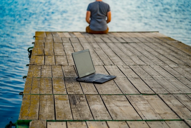Junger Mann, der auf einem Pier sitzt und seinen Laptop benutzt. Arbeit und Urlaub.