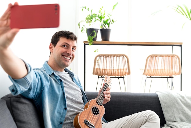 Junger Mann, der auf einem Sofa sitzt und mit seinem Handy ein Selbstporträt macht, während er Ukulele spielt