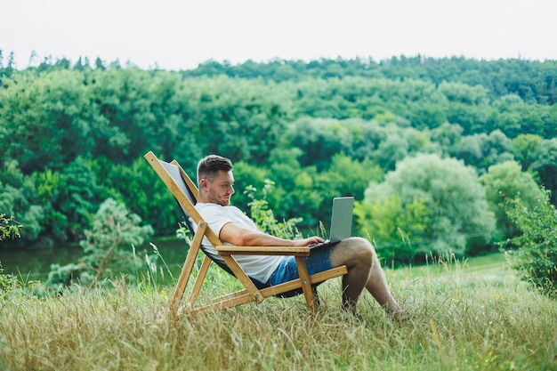 Junger Mann, der auf einem Stuhl in der Natur liegt und mit einem Laptop arbeitet, der allein ruht und nachdenklich nachschaut. Ein Mann auf einer Reise auf dem Land, das Konzept des Lebensstils der Menschen