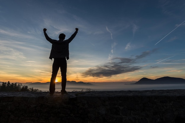 Junger Mann, der auf einer alten Mauer unter herrlichem Abendhimmel steht und seine Arme erfolgreich hoch erhoben hat.