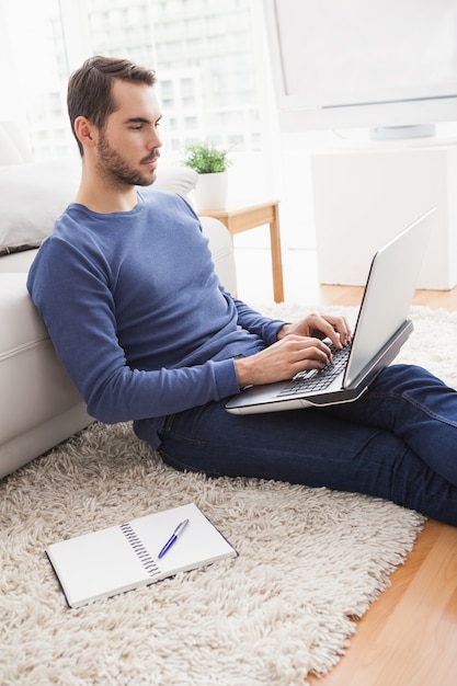 Junger Mann, der auf Fußboden unter Verwendung des Laptops sitzt