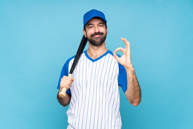 Junger Mann, der Baseball über der lokalisierten blauen Wand zeigt okayzeichen mit den Fingern spielt