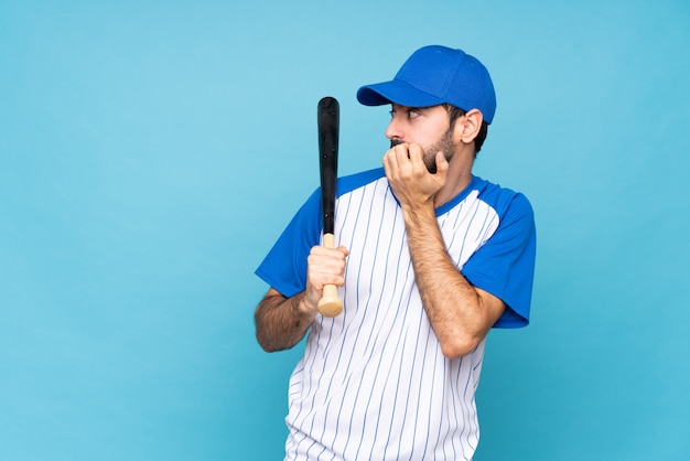 Junger Mann, der Baseball über lokalisierter blauer Wand nervös und erschrocken spielt, Hände zum Mund setzend