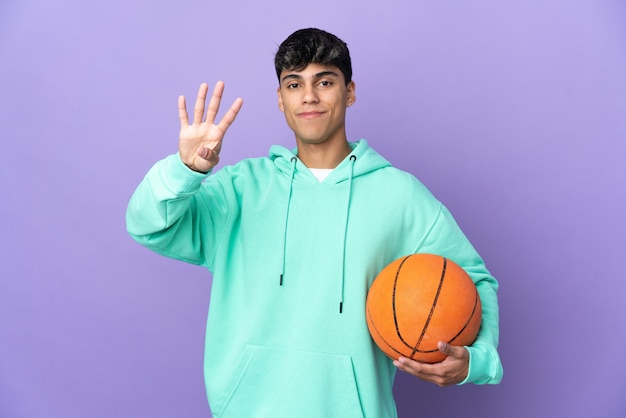 Junger Mann, der Basketball über lokalisierten lila Hintergrund glücklich spielt und vier mit den Fingern zählt