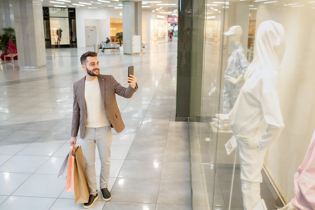 Junger Mann, der beim Einkaufen im Einkaufszentrum ein Foto von Mannequin am Telefon hinter dem Fenster macht