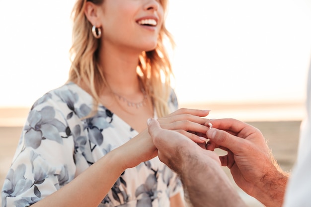 Junger Mann, der beim Gehen am sonnigen Strand einen Verlobungsring an den Finger der Frau legt