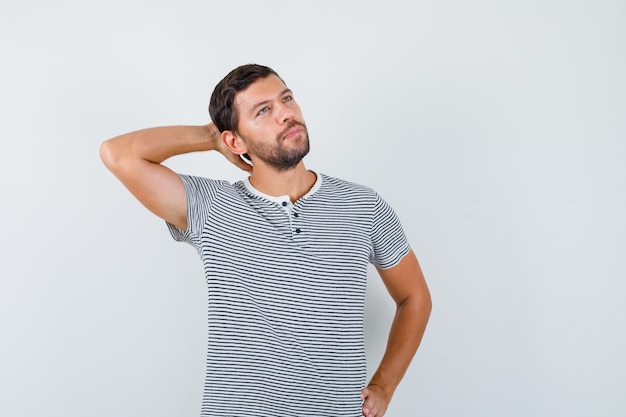 Junger Mann, der die Hand im T-Shirt hinter dem Kopf hält und nachdenklich aussieht. Vorderansicht.