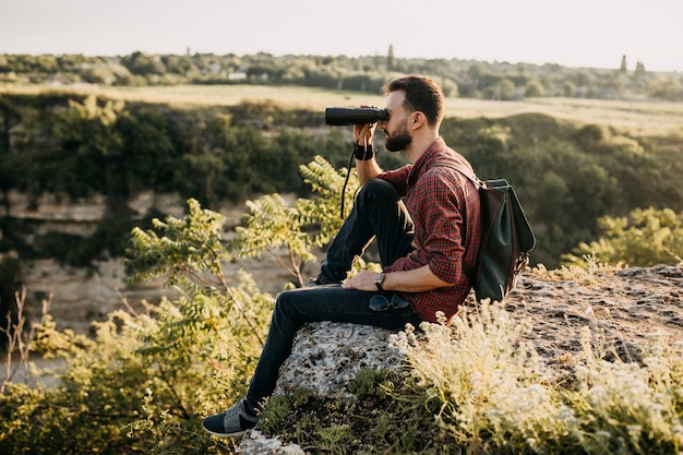 Junger Mann, der durch Fernglas schaut und auf einer Klippe in den Bergen sitzt.