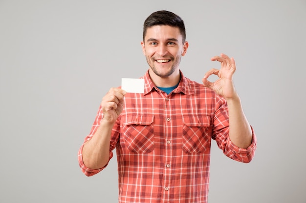 Junger Mann, der eine leere Visitenkarte auf grauem Hintergrund hält und zeigt