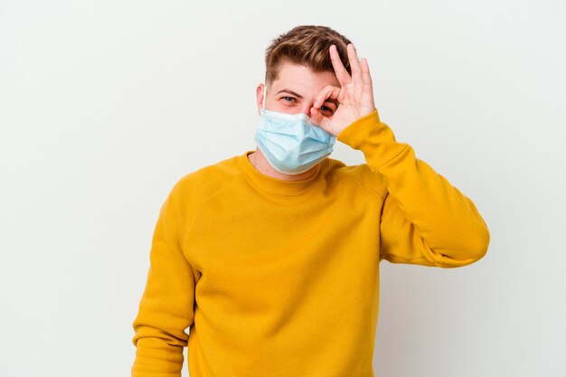 Junger Mann, der eine Maske für Coronavirus trägt, lokalisiert auf weißer Wand aufgeregt, hält ok Geste auf Auge.