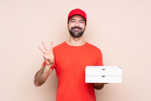 Junger Mann, der eine Pizza über lokalisiertem Hintergrund glücklich hält und drei mit den Fingern zählt