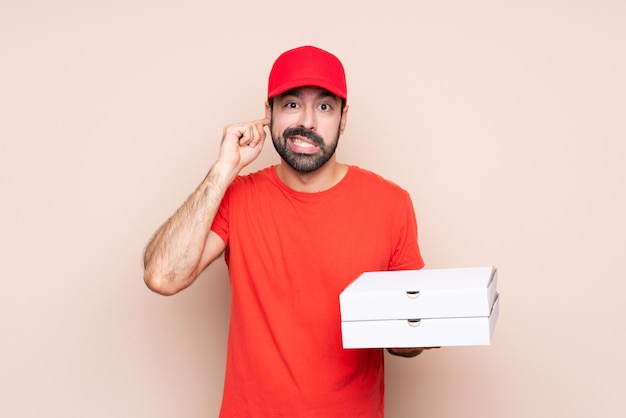 Junger Mann, der eine Pizza über lokalisierten frustrierten und Bedeckungsohren hält