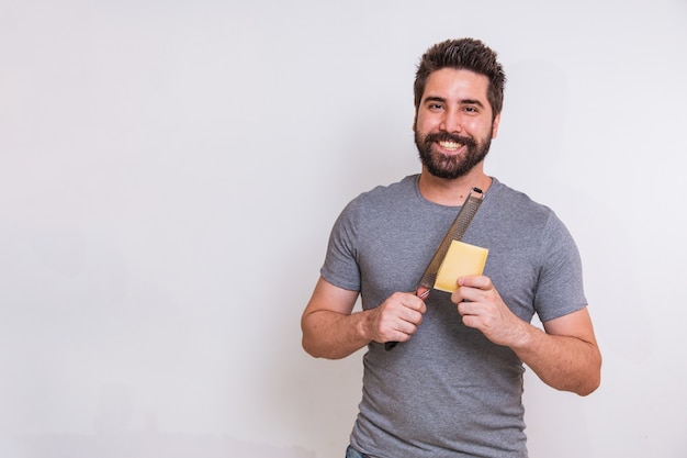 Junger Mann, der eine Reibe und ein Stück Käse in der Hand auf grauem Hintergrund hält. Mann wird den Käse reiben