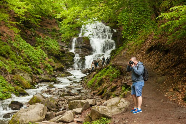 Junger Mann, der eine schwarze Kamera hält und Fotos auf dem Hintergrund des Wasserfalls im Wald macht