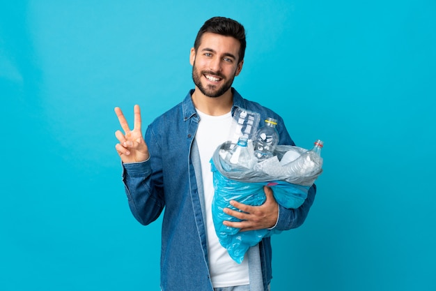 Junger Mann, der eine Tasche voller Plastikflaschen hält, um lokalisiert auf der blauen Wand zu recyceln, die lächelt und Siegeszeichen zeigt