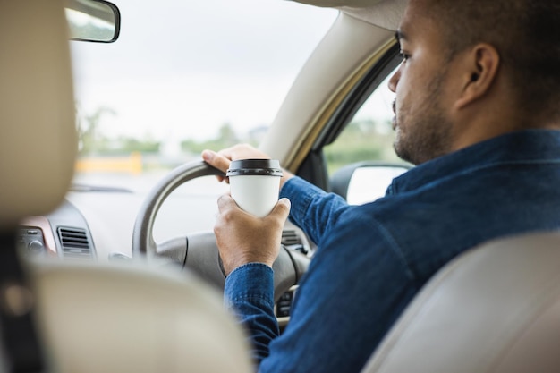 Junger Mann, der eine Tasse heißen Kaffee trinkt, während er mit dem Auto fährt, um zu reisen Hände mit Lenkrad