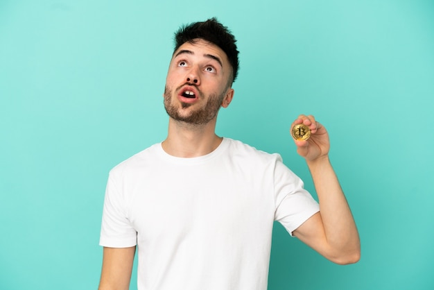 Junger Mann, der einen Bitcoin auf blauem Hintergrund isoliert hält und nach oben schaut und mit überraschtem Ausdruck