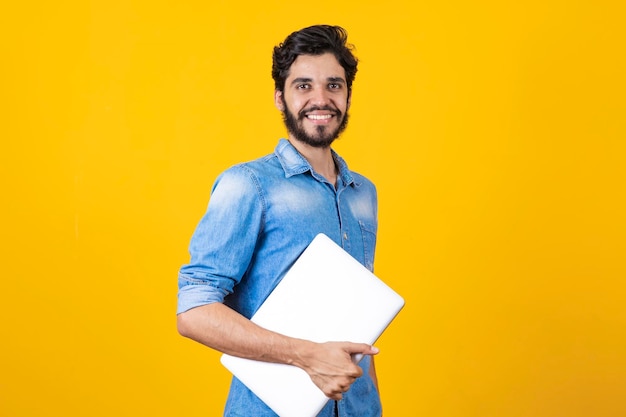 Junger Mann, der einen Computer hält, schloss in seinen Armen auf gelbem Hintergrund Geschäftsmannkonzept