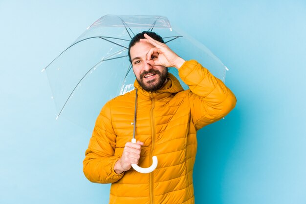 Junger Mann, der einen langen Haarblick hält, der einen Regenschirm aufgeregt hält, ok Ordnung Geste auf Auge haltend.