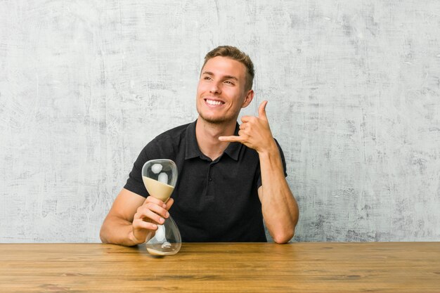 Junger Mann, der einen Sandtimer auf einer Tabelle zeigt eine Handyanrufgeste mit den Fingern hält.