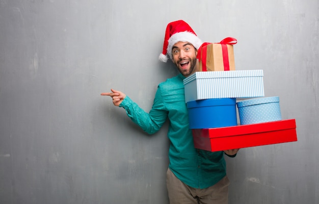 Junger Mann, der einen Sankt-Hut hält die Geschenke zeigt auf die Seite mit dem Finger trägt